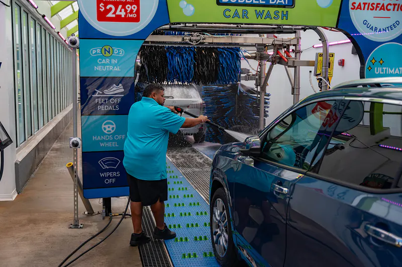 A team member pressure-washing the front of a blue SUV before it enters the tunnel